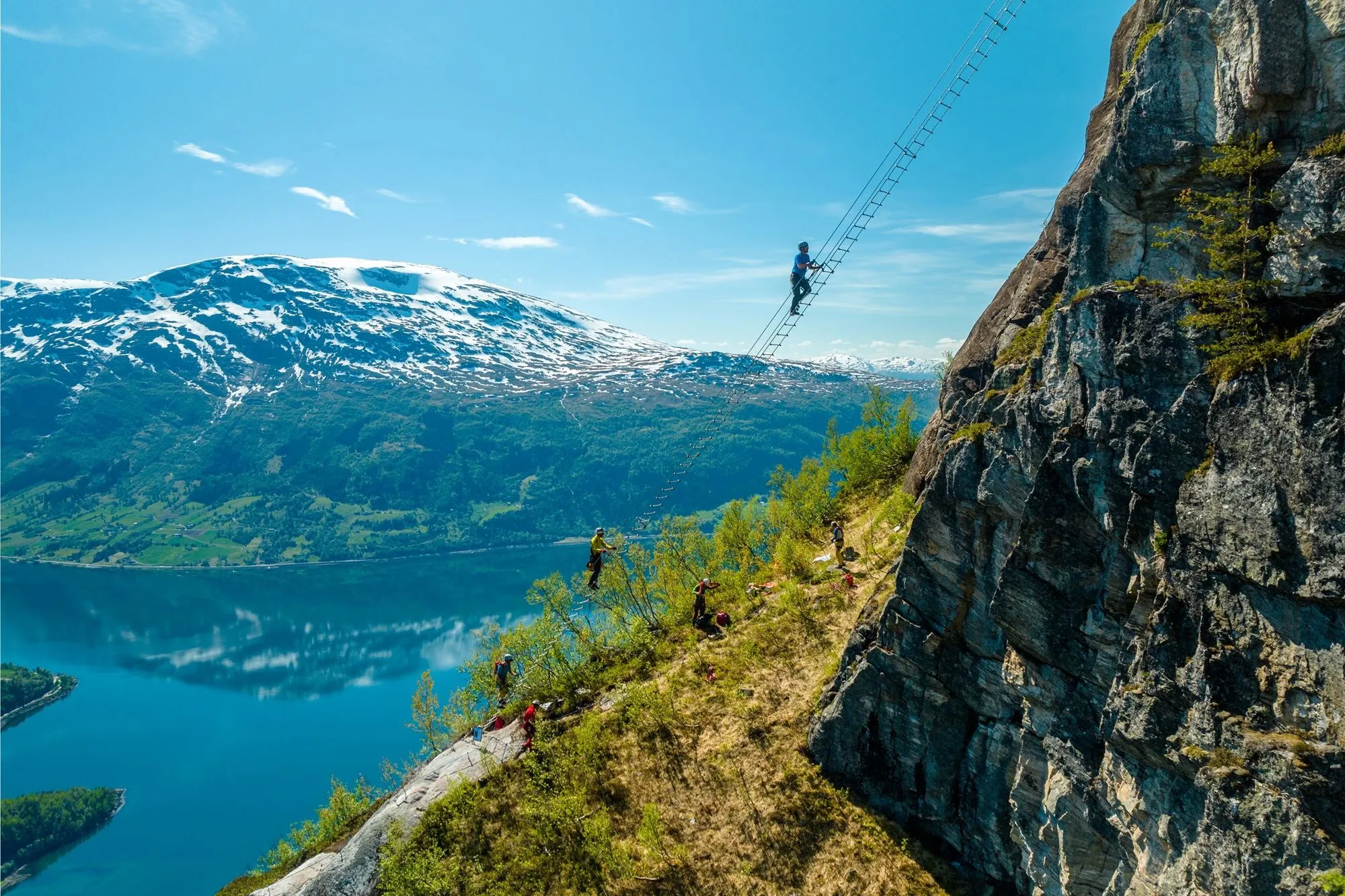 Ein person klatrar på ei luftig stige ved ein bratt fjellvegg, med fjord og snødekte fjell i bakgrunnen..