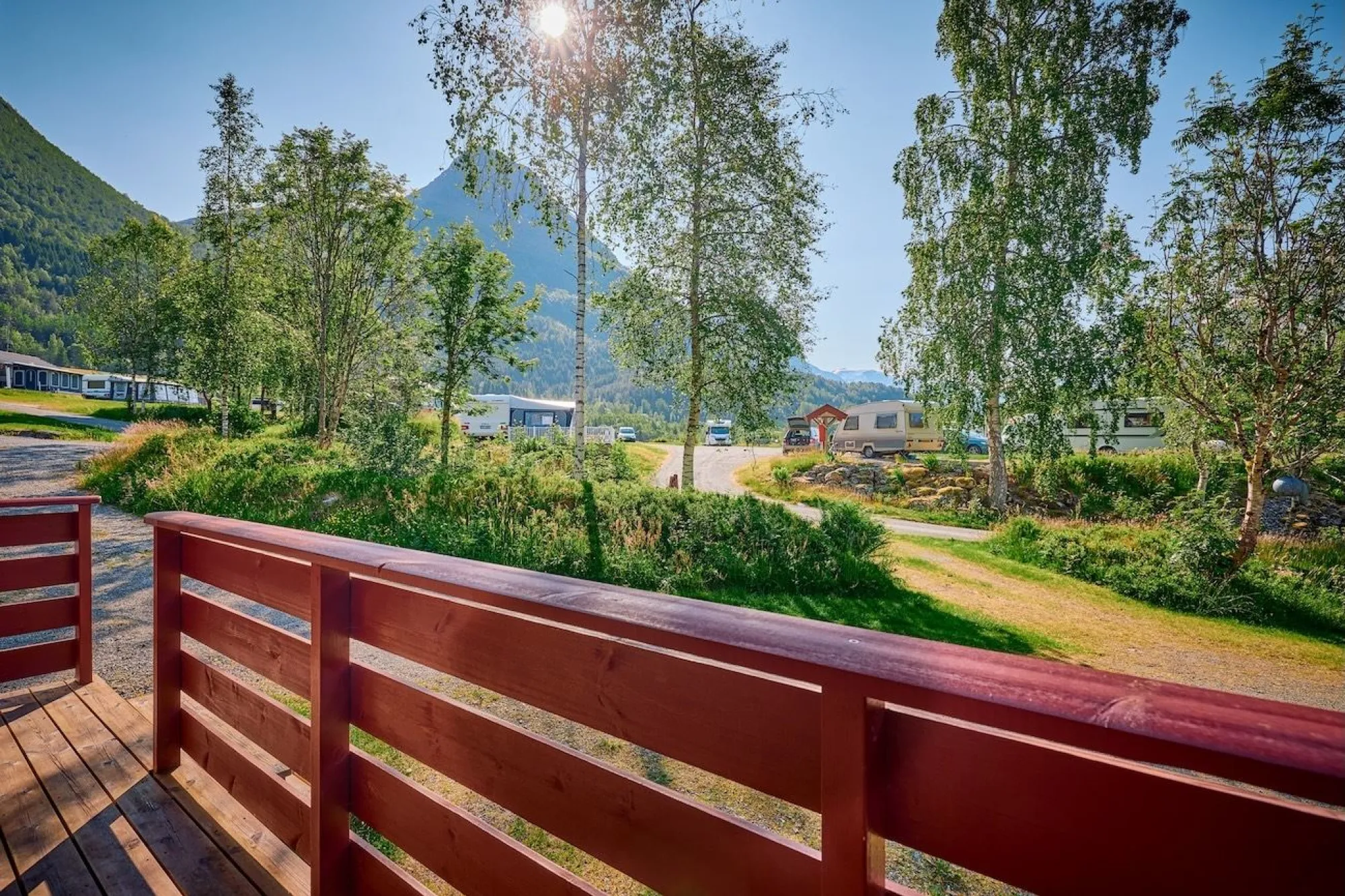 Red wooden deck railing overlooks a green campground with trees, caravans, and mountains under bright sun..