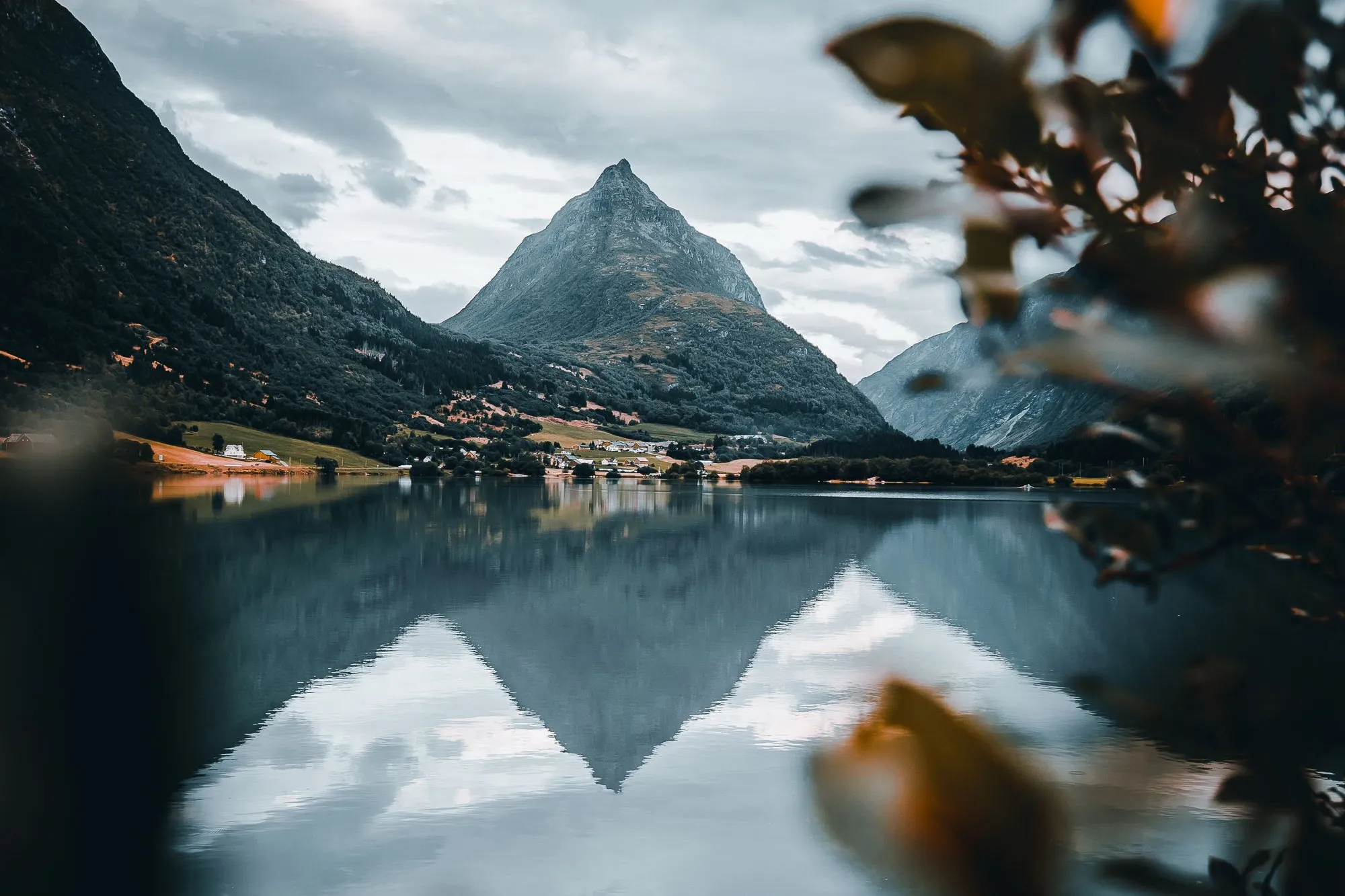 Spiss fjelltopp ved ein stille fjord med klår spegling, små bygder langs stranda og skyer på himmelen..