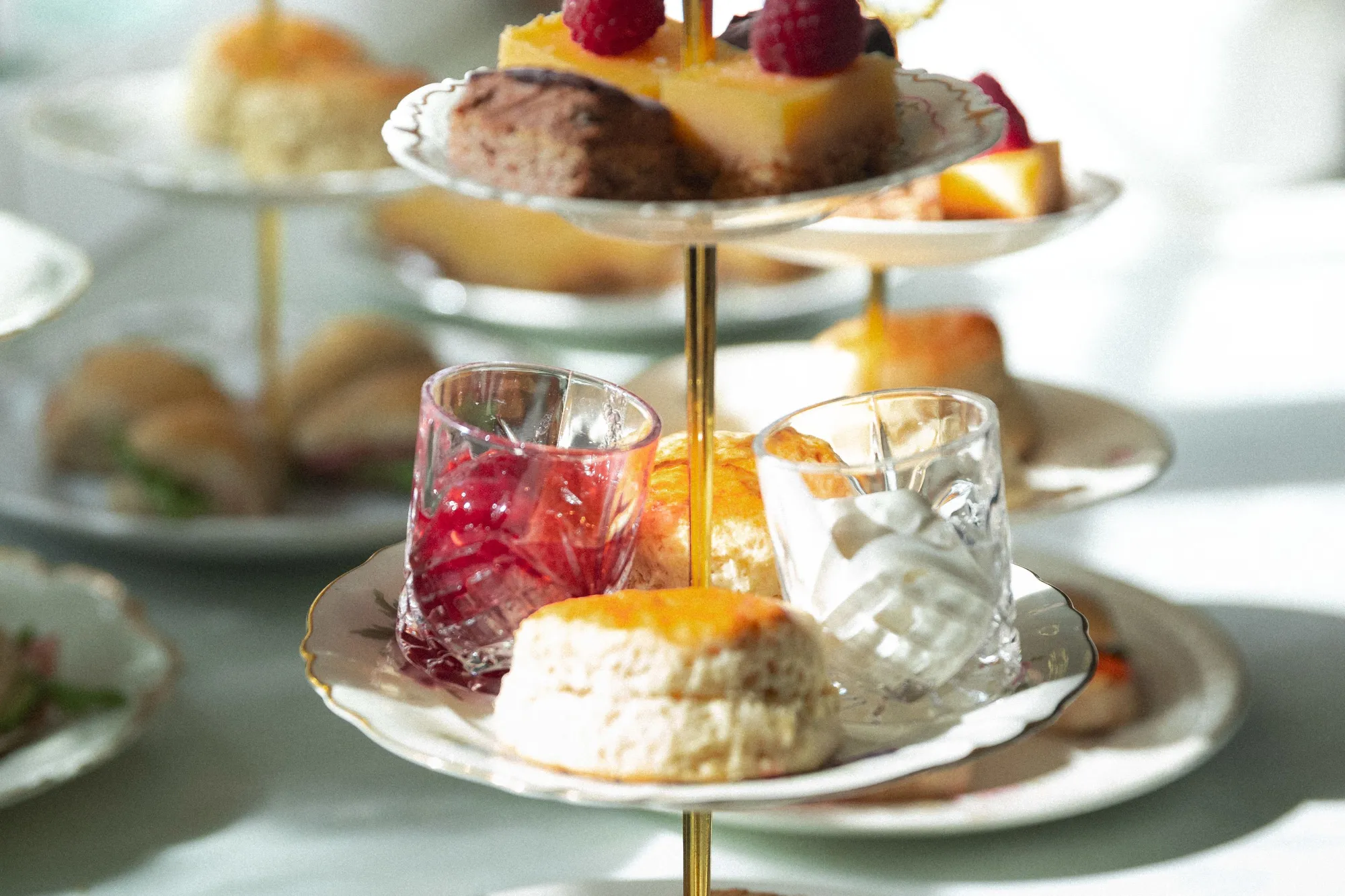 Eit etasjefat med små smørbrød, scones, syltetøy og kaker med bringebær, i mjukt dagslys..