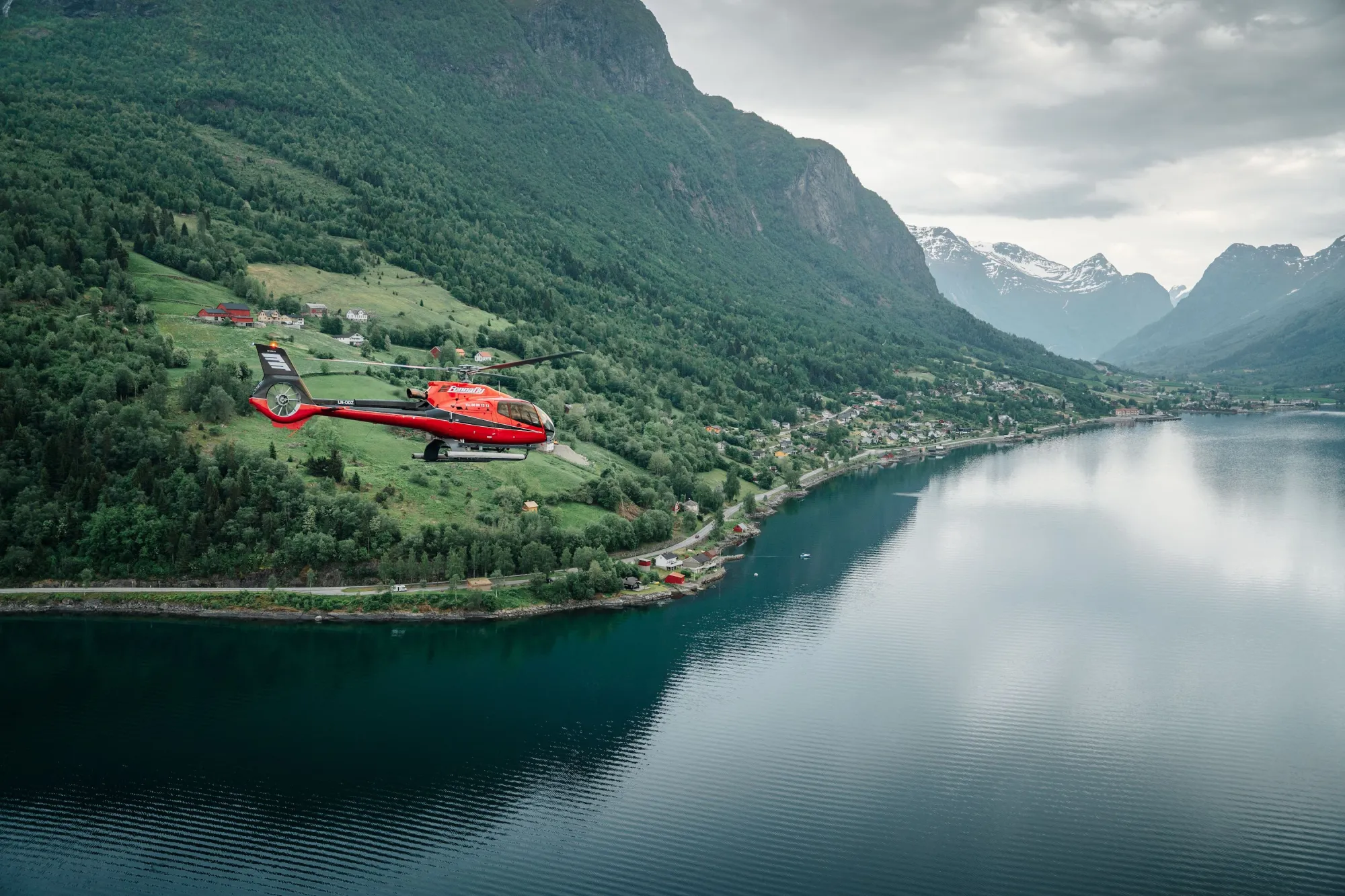 Fjord Helikopter - Eit raudt helikopter flyg over ein fjord med skogkledde fjell, bygder langs stranda og skya himmel..