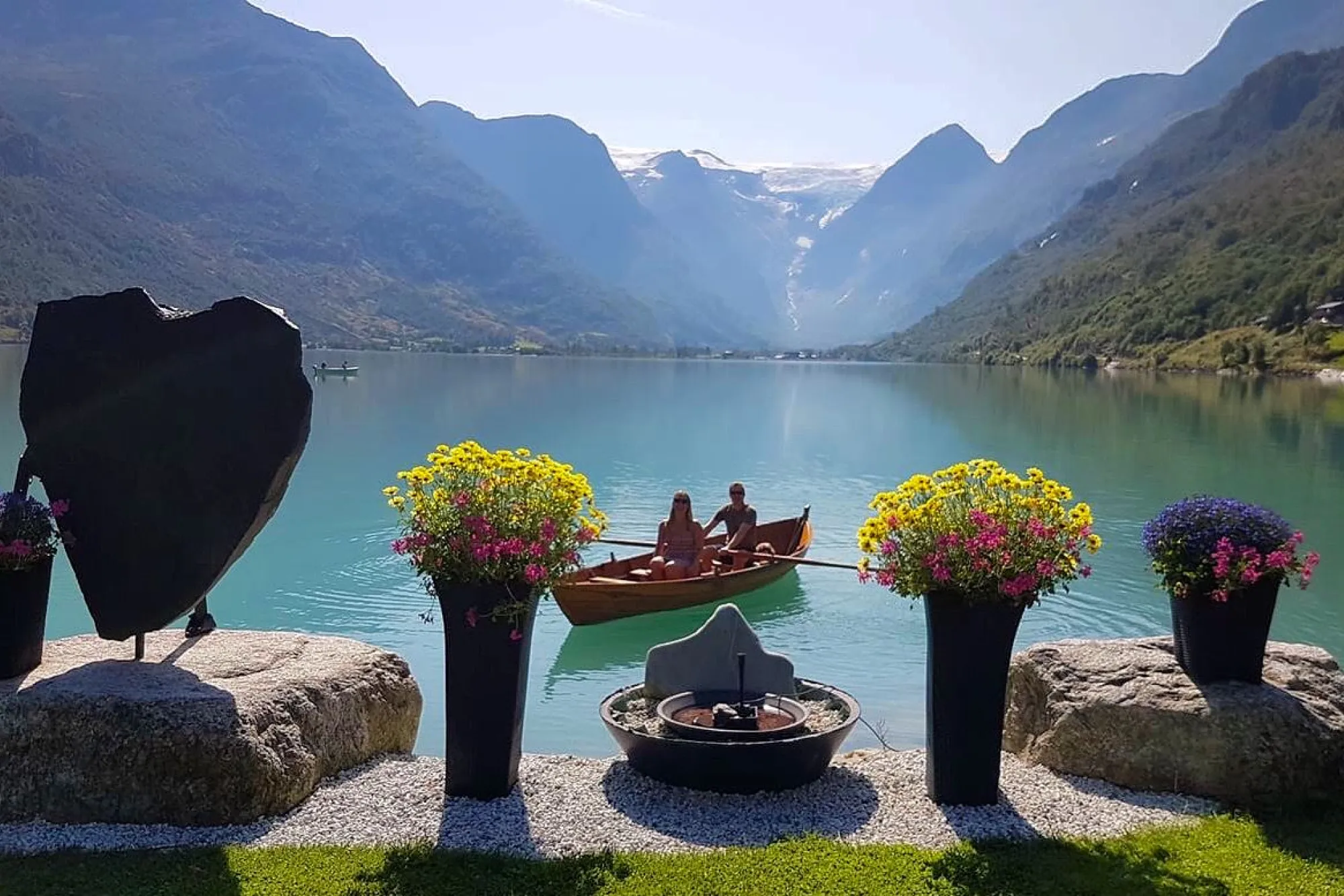To personar i ei lita trebåt på turkis fjordvatn, med fjell og bre i bakgrunnen og blomepotter i framgrunnen..