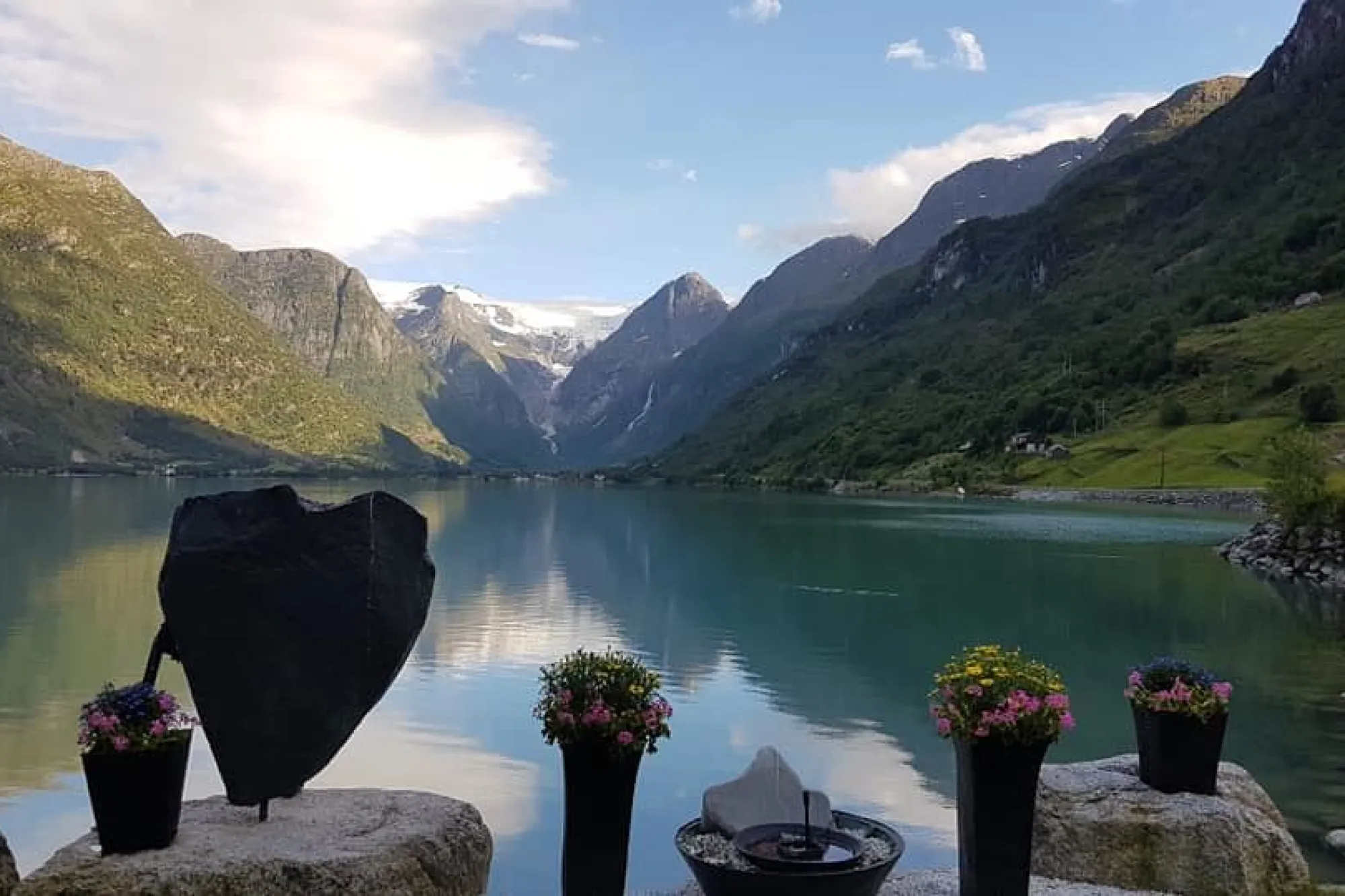 Stille Oldenvatnet med spegling av fjell og skyer, sett frå steinplatå med blomsterpotter og ei hjartforma skulptur på Yrineset.