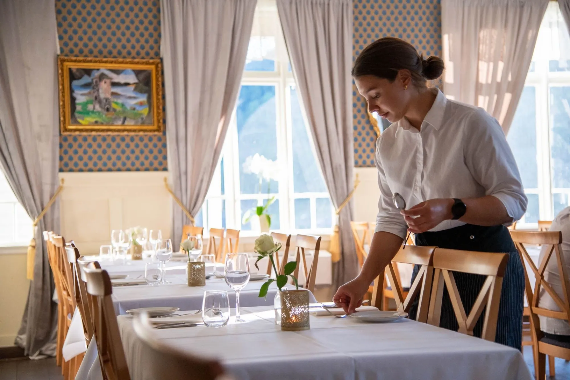 Ein servitør i kvit skjorte dekkjer eit bord med kvit duk, glas og blome, i ein lys restaurant med vindauge..
