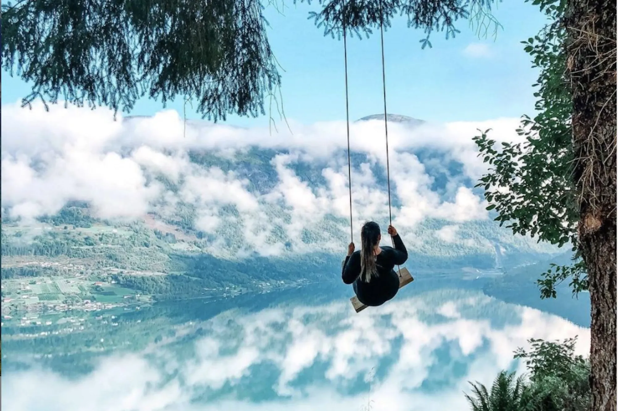 Ein person sit på ei huske som heng i eit tre, med fjord, fjell og skyer i bakgrunnen..