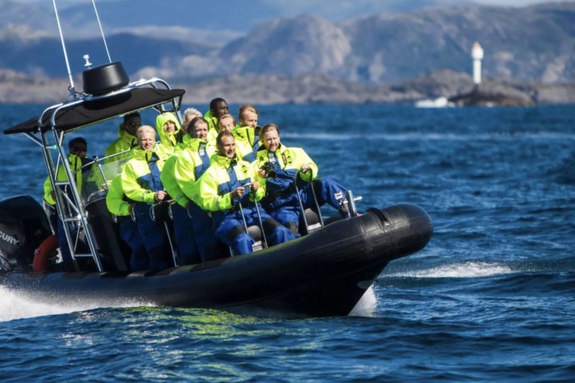 Svart RIB-båt med fleire personar i gule jakker køyrer fort på havet, med fjell og fyr i bakgrunnen..