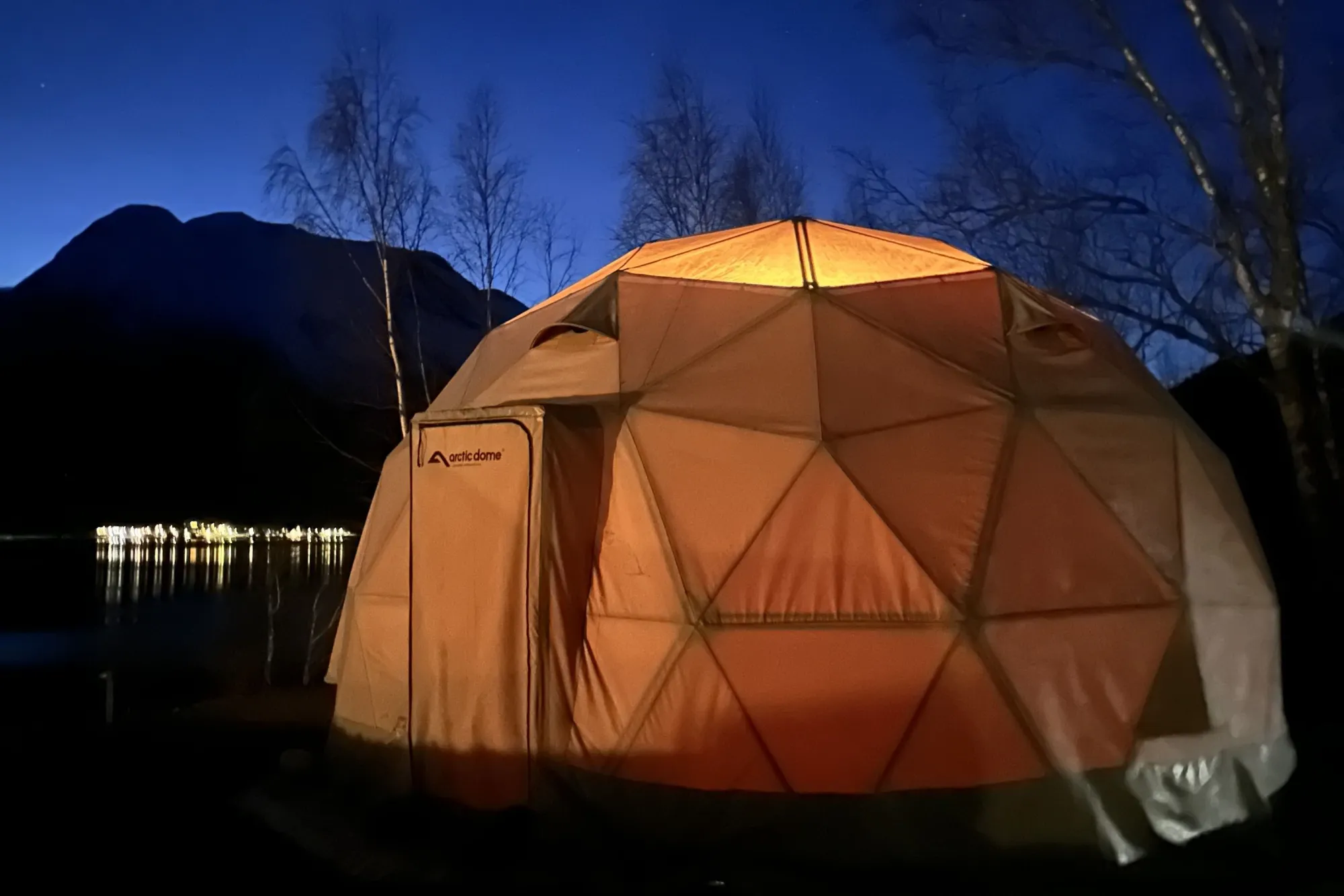 Eit opplyst oransje kuppeltelt står ved ei mørk strand, med fjell, nakne tre og stjernehimmel bak på Vollsnes i Oppstryn ved Oppstrynsvatnet..
