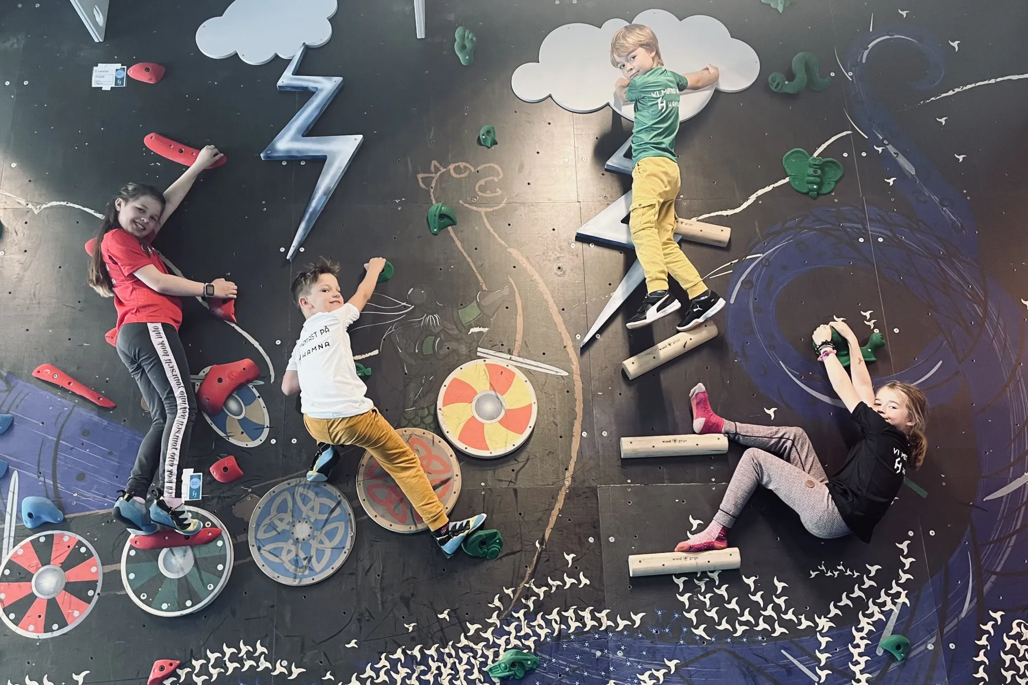Fire barn klatrar på ei innandørs klatrevegg med fargerike tak, teikningar av skyer og viking-skjold..