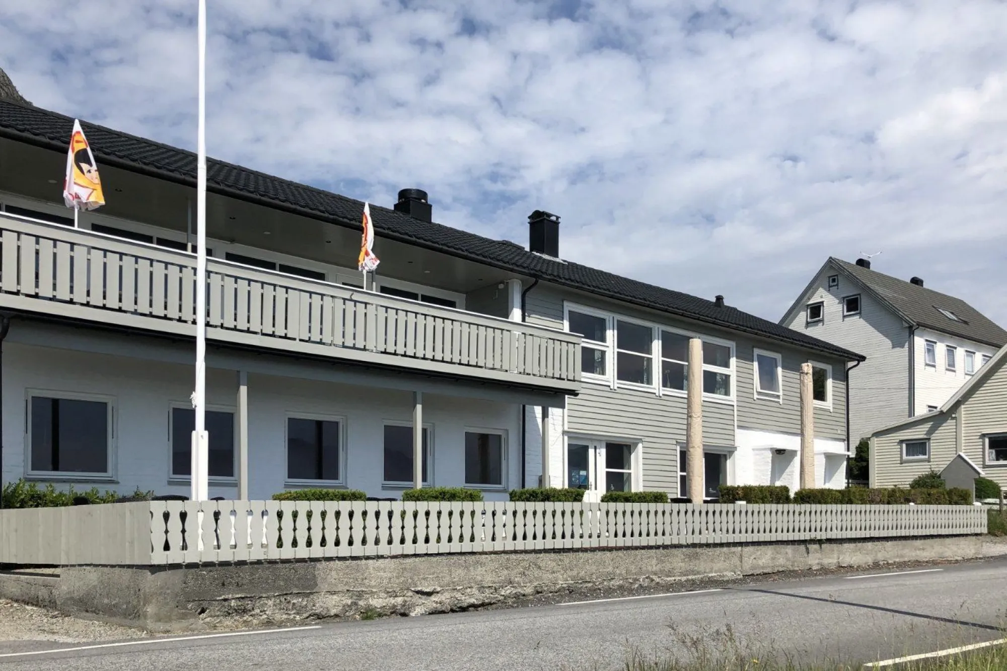 Eit grått, toetasjars bygg med balkong, flaggstong og kvitt gjerde langs ei veg under skya himmel..