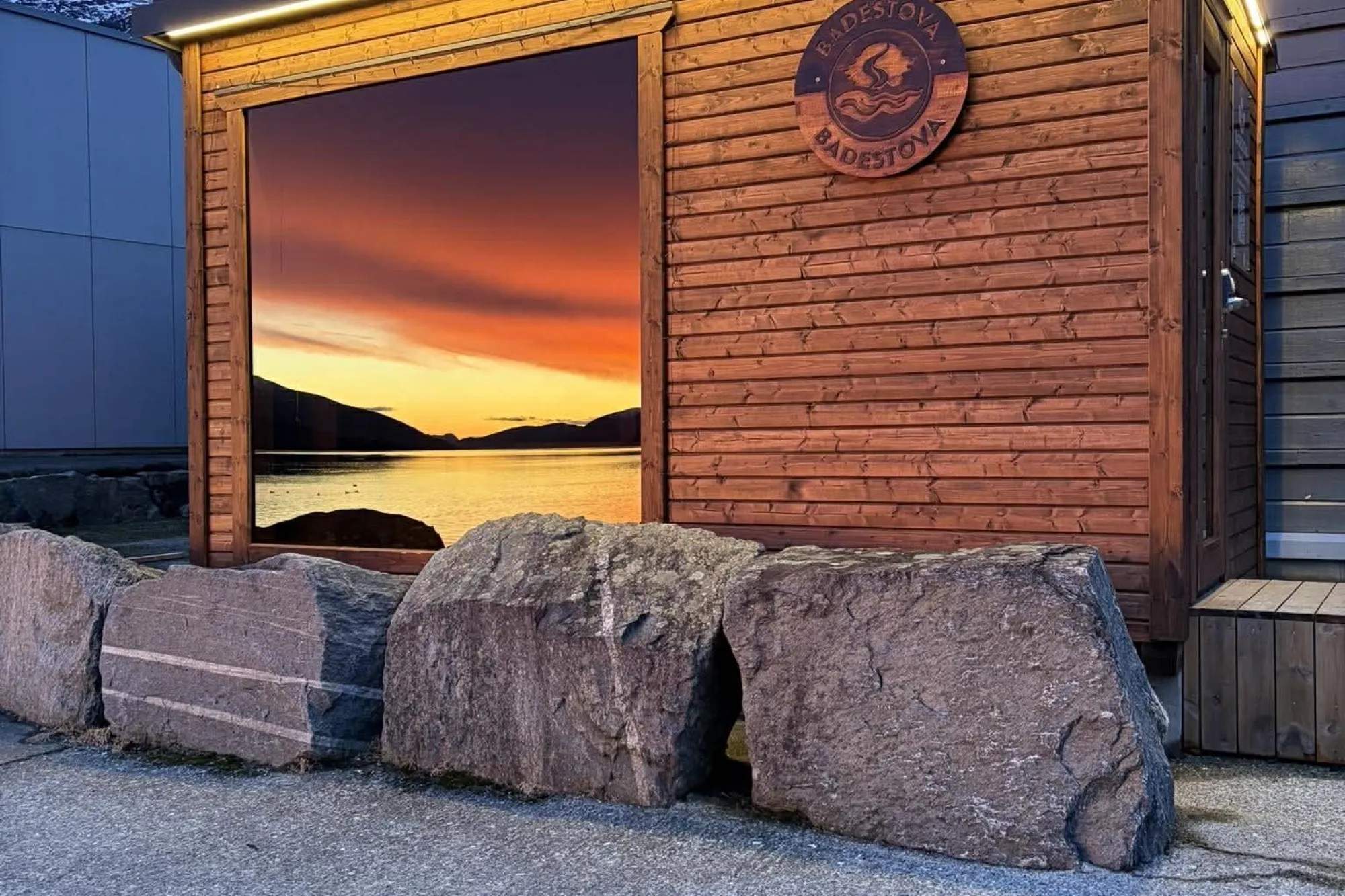 Badstova Sauna med lyslist og stort bilete av solnedgang over vatn og fjell; store steinblokker framfor..